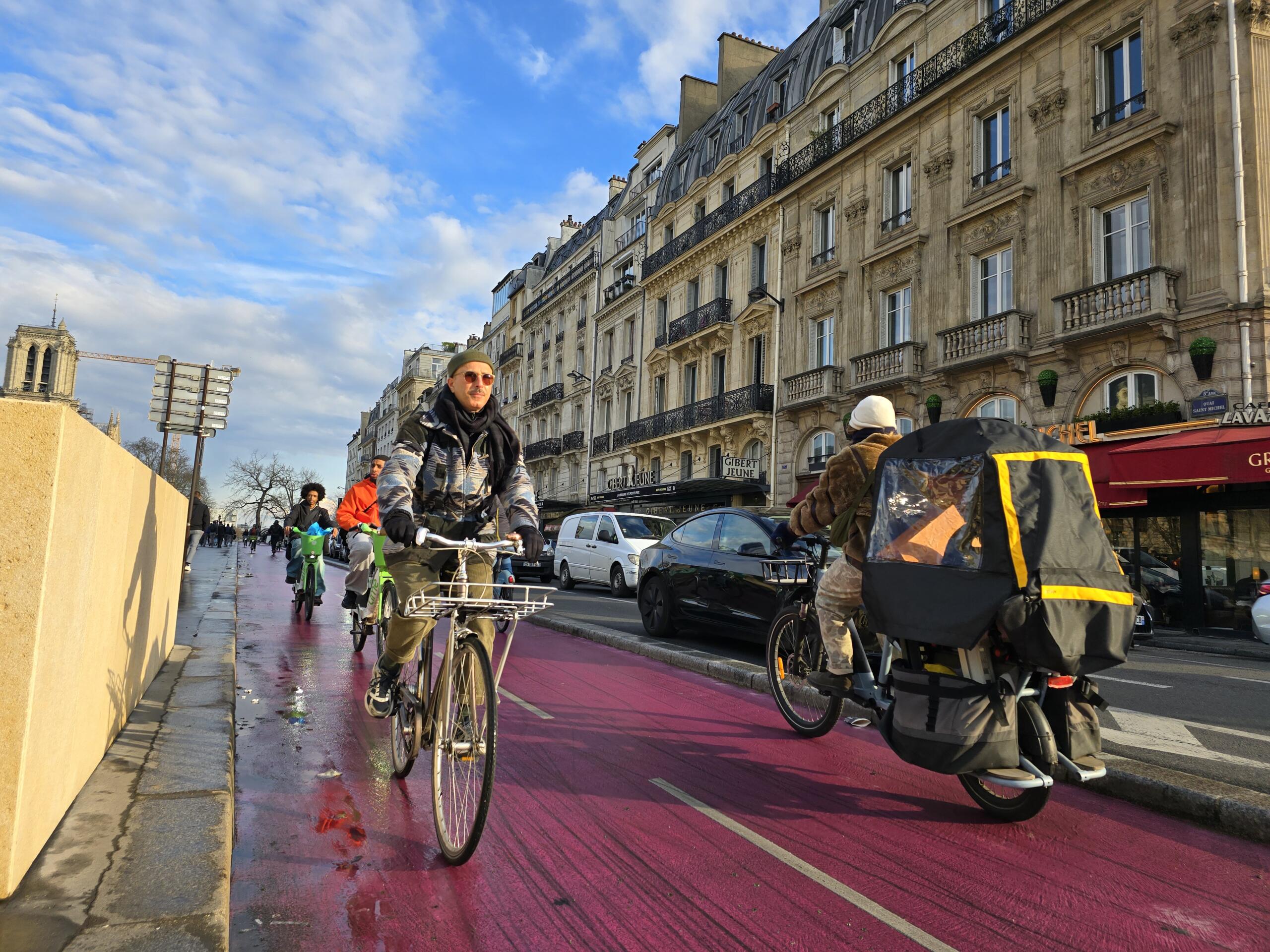 Photo of a bike lane in a city. There are a man on a bike, a woman carrying a kid in the cargo bike, and behind them a young woman and a young man riding green shared bikes.