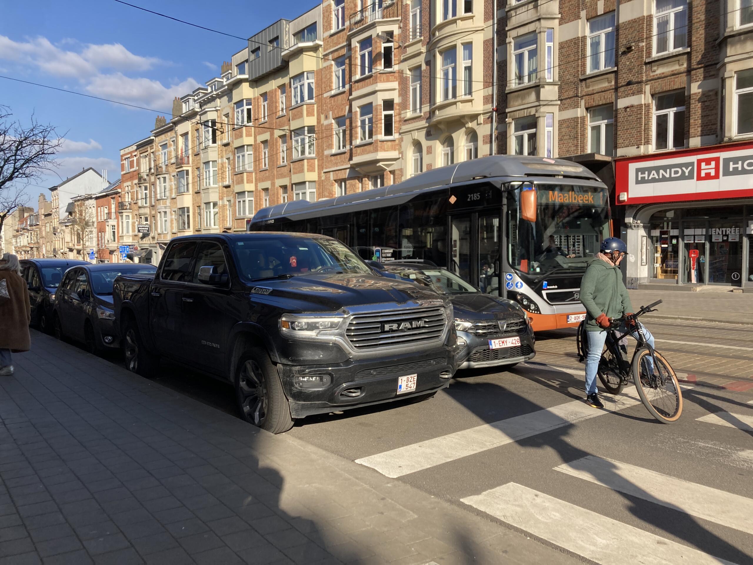 Photo of vehicles waiting at a traffic light. On the left, a black SUV towers over a black car. In front a man riding a bike and a bus