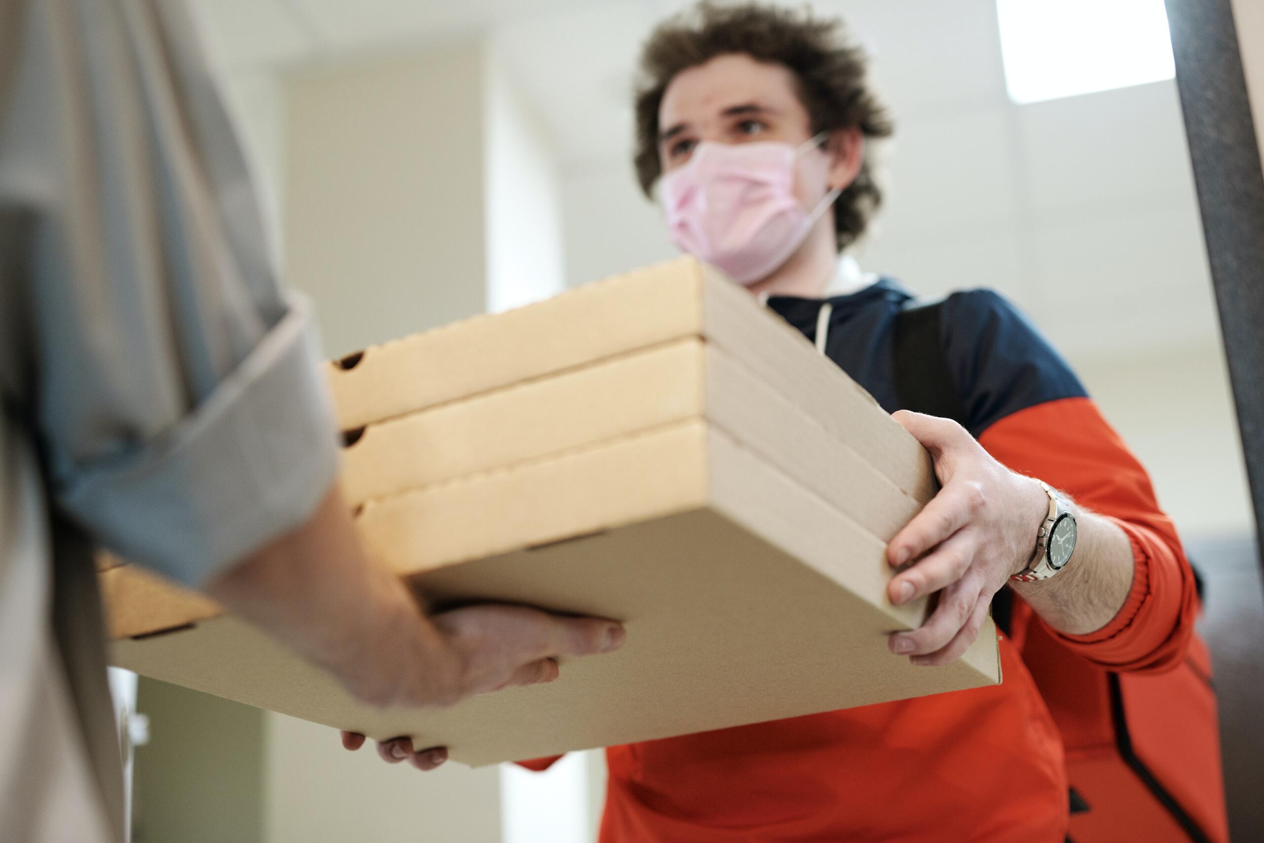 Photo of a young man wearing a face mask and a red and black sweater handing pizza boxes to another person