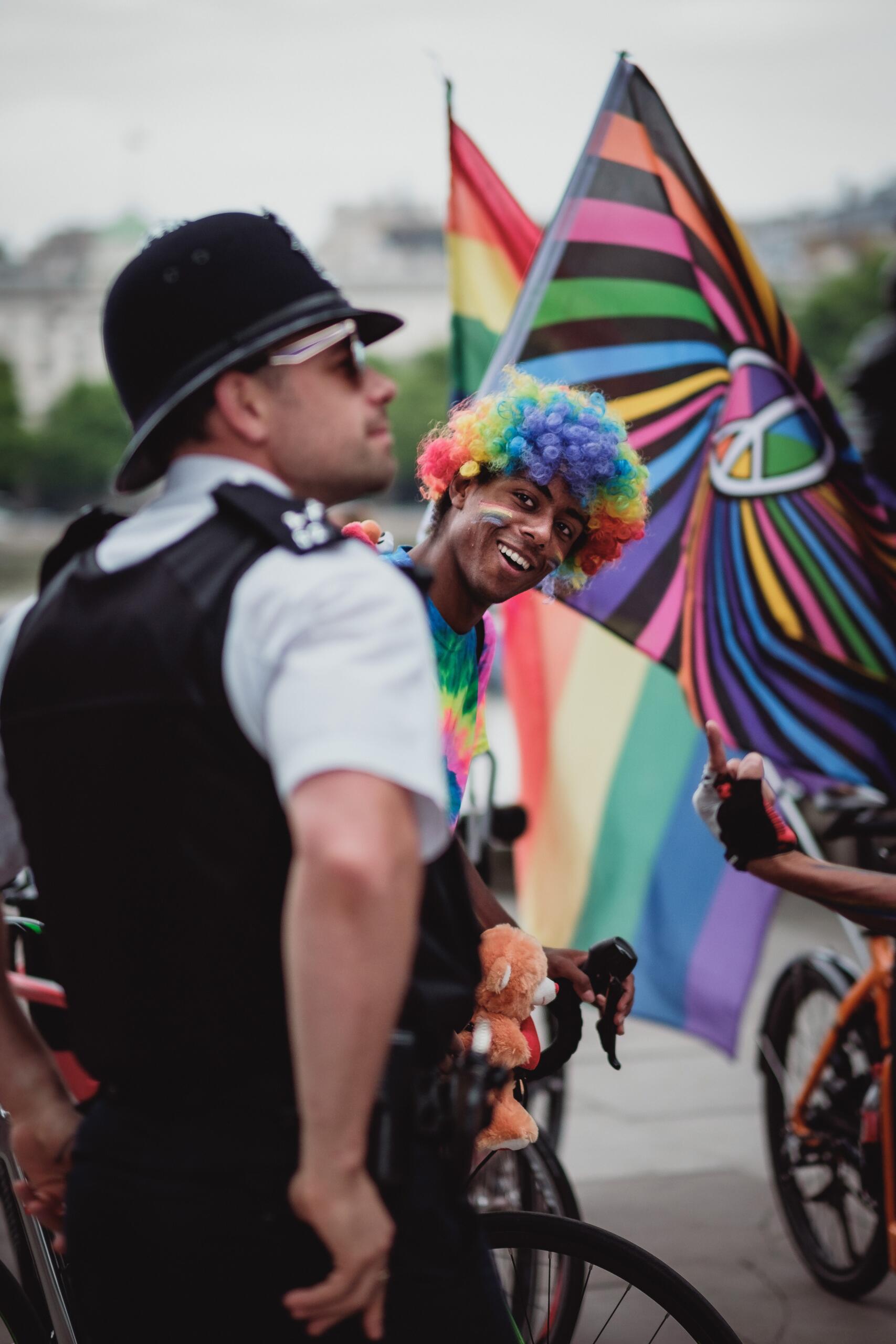 Photo of a police man and behind him a person wearing a rainbow wig peaking at the camera