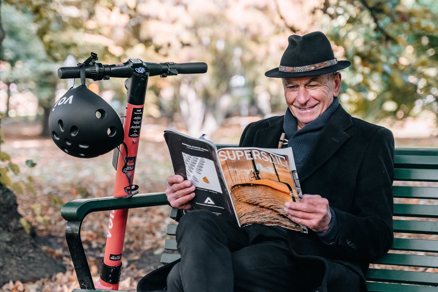 Photo of an old man sitting on a bench while reading a magazine with a Voi scooter parked next to him