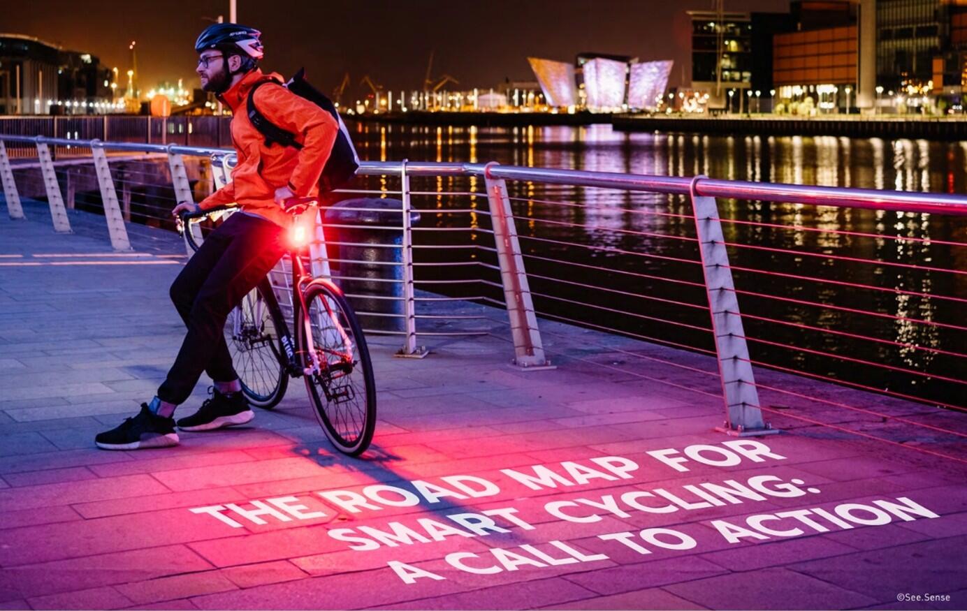 Photo of a cycling leaning on his back, which has a light projector attached showing the text 'The road map for smart cycling: a call to action" on the pavement